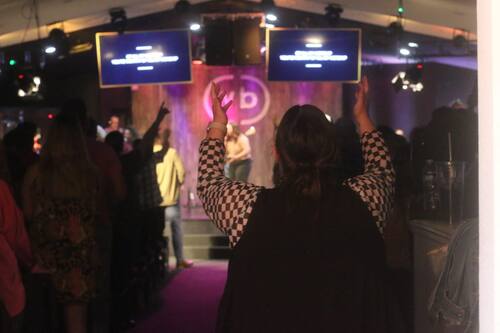 View from the back of the WBCC sanctuary during a Sunday service, showing a congregant with hands raised in worship, surrounded by others also worshiping with hands lifted. The scene captures a powerful moment of collective praise.