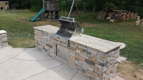 Outdoor kitchen area with a built-in grill and stone countertop, set in a backyard with a playground in background.