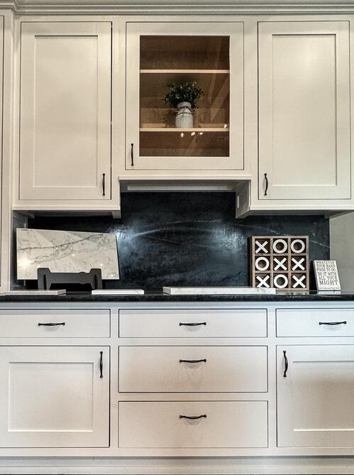 White kitchen cabinets with black handles, featuring a glass-front cabinet displaying a potted plant, black countertop, and decorative items including a marble slab.