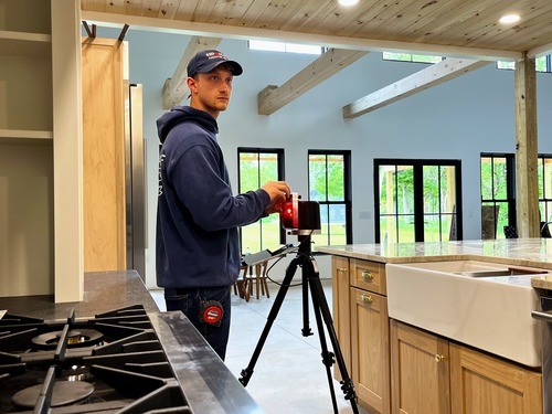 Red Leaf Milling Company staff member wearing a navy blue hoodie and hat, operating a camera on a tripod in a kitchen with custom wood cabinetry and a farmhouse sink