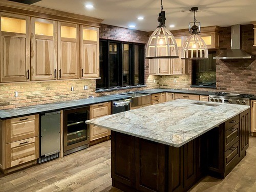 Spacious kitchen featuring custom light wood cabinets with black handles, a large marble-topped island, built-in stainless steel appliances, and pendant lighting. The kitchen also has a brick backsplash and wood flooring.