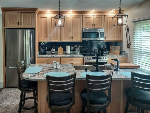 Kitchen with light wood cabinets, stainless steel appliances, black subway tile backsplash, marble countertops, and a kitchen island with bar seating and pendant lighting.
