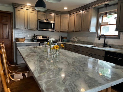 Close-up of a kitchen island with a marble countertop, grey cabinets, black appliances, and pendant lighting, featuring small vases with flowers as a centerpiece.