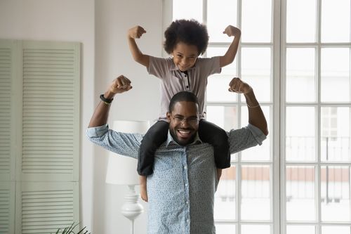 Small son sit on strong dad shoulders showing biceps. African family enjoy activity games at home, healthy fit lifestyle, two superheroes, vitamins for adults and children ad, happy Father Day concept. Credit: fizkes 