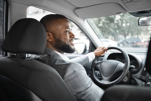 Cheerful african american businessman in silver suit driving car, shot from behind. Happy bearded black man driver holding hands on wheel and gear, driving luxury car, looking at empty space