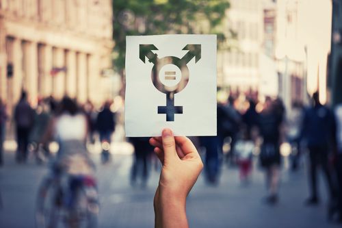  Hand holding a paper sheet with transgender symbol and equal sign inside. Equality between genders concept over a crowded city street background. Sex rights as a metaphor of social issue.