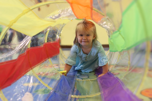 Little girl in blue dress crawling through rainbow colored tube