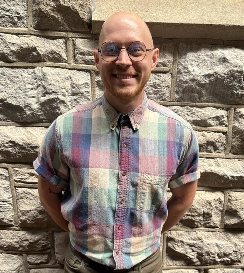 An image of Fred Mottaz, salt and pepper hair with glasses, wearing a white and blue checkered button up shirt with a big smile in front of the stones in the atrium of First Christian.