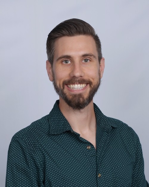 An image of Rev. Dr. Brad Stagg, bald-headed with nicely framed wire glasses, wearing a blue suit & sweater with a big smile in front of the stones in the atrium of First Christian.