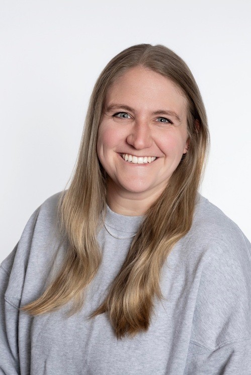An image of Tracy Westhoff, dark brown hair and a few light freckles, wearing a white shirt and black trim with a big, pink-lipsticked smile in front of the stones in the atrium of First Christian.