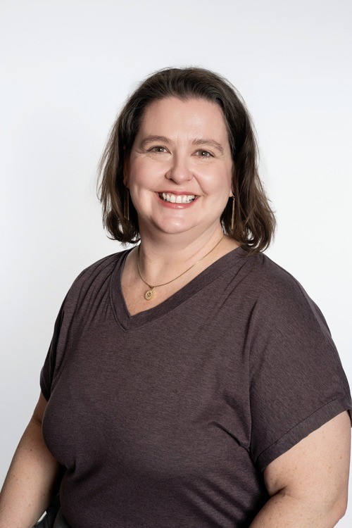 An image of Tracy Westhoff, dark brown hair and a few light freckles, wearing a white shirt and black trim with a big, pink-lipsticked smile in front of the stones in the atrium of First Christian.