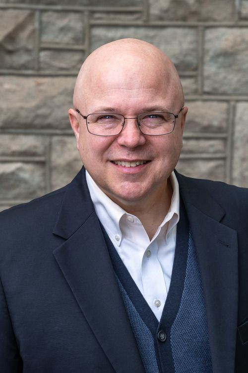 An image of Rev. Dr. Brad Stagg, bald-headed with nicely framed wire glasses, wearing a blue suit & sweater with a big smile in front of the stones in the atrium of First Christian.