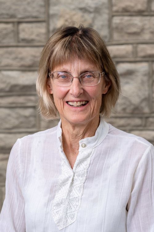 An image of Dr. Rochelle Parker, blonde and brown hair with glasses, wearing a white blouse with a big smile in front of the stones in the atrium of First Christian.