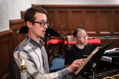 An image of Michael Sauer directing music from a hymnal on a music stand on the sanctuary stage, as Travis McFarlane plays the piano in the background.
