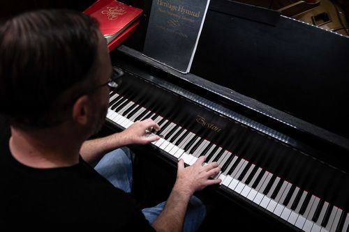 An image of Travis McFarlane playing the piano in the sanctuary of the church, wearing a black shirt and blue jeans.