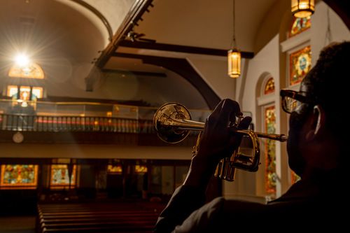 An image of Carlot Dorve playing a trumpet with one hand fancing the sanctuary as the sun shines beautifully through the stain glass windows of the balcony.