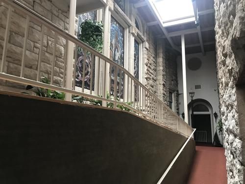 Image of the ramp in the atrium of First Christian Church, red carpet with a white railing and clean, beige stone lit well by skylights. On the left are large stain glass windows, slightly covered by lush green houseplants.