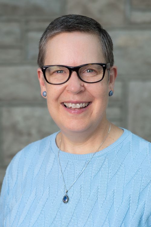 An image of Rev. Sally Robinson, darker blond hair with sprinkles of gray and dark framed glasses, wearing a blue sweater with a big smile in front of the stones in the atrium of First Christian.