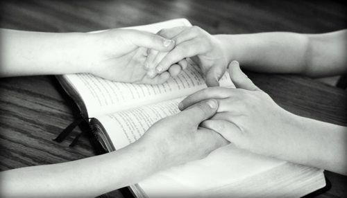 Image of two people holding hands in prayer with a Bible underneath their hands