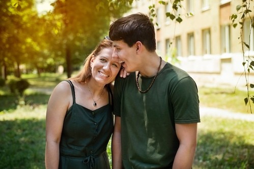 A young man gently kisses a woman on the head as they smile, enjoying a warm moment together outside.