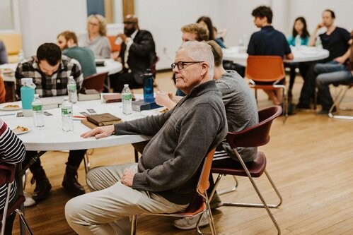 groups of people at circle tables in a classroom, elderly man in center