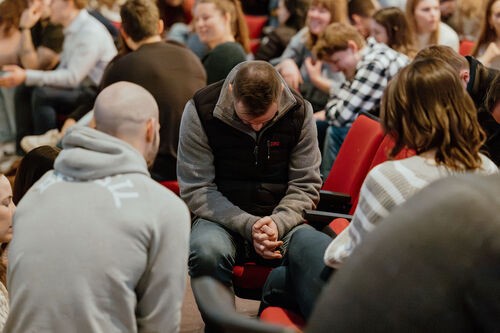 A group of people sitting in prayer, the man in the center has his head bowed and hands folded