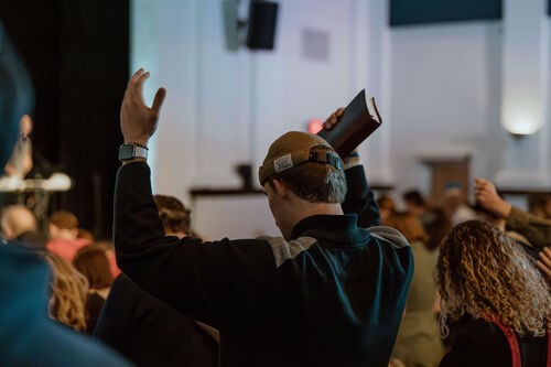 A young man in a congregation with his arms lifted in worship and a Bible in his right hand