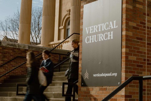 a few people walking down the front steps of Vertical Church with a large sign to their right that reads,